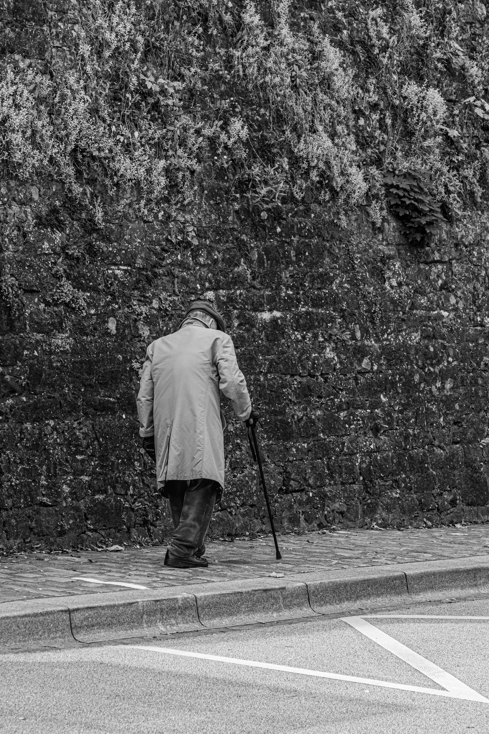 Photographie d'art noir et blanc d'un vieil homme avec une canne devant un mur de pierre - Tirage unique Masterpiece.